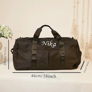 May include: Black duffel bag with the name "Nika" embroidered in white. The bag is 18 inches long, 10.2 inches high, and 7.8 inches wide. It has two handles and a shoulder strap. The bag is on a white surface.