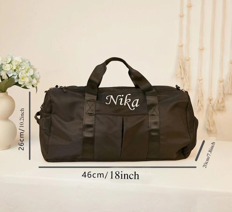 May include: Black duffel bag with the name "Nika" embroidered in white. The bag is 18 inches long, 10.2 inches high, and 7.8 inches wide. It has two handles and a shoulder strap. The bag is on a white surface.