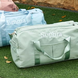 Puede incluir: Tres bolsas de lona personalizadas en colores pastel: verde claro, azul claro y rosa. El nombre "Sophia" está bordado en blanco en la bolsa verde y "Emma" en la bolsa azul. Las bolsas tienen asas, cremalleras y bolsillos.