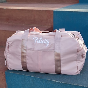 May include: A blush pink duffel bag with the name "Felicity" embroidered in white script. The bag has a rectangular shape with a zipper closure, two handles, and a shoulder strap. The bag is sitting on a blue surface.