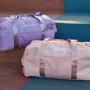 May include: Two personalized duffel bags. One is lavender with the name "Janelle" embroidered in white script. The other is a light pink with "Felicity" embroidered in white script. Both bags have matching straps and zippers.