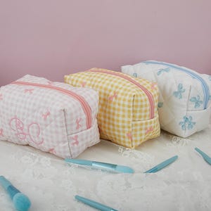 May include: Three quilted makeup bags with gingham patterns and embroidered bows. The bags are in pink, yellow, and white.