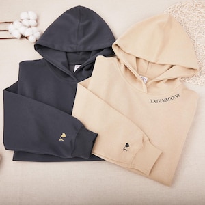 Op de afbeelding: Twee hoodies, één in antracietgrijs en één in beige, worden getoond. De grijze hoodie heeft een gouden hart en de letter "Y" geborduurd op de mouw. De beige hoodie heeft een zwart hart en de letter "T" geborduurd op de mouw, met Romeinse cijfers op de capuchon.