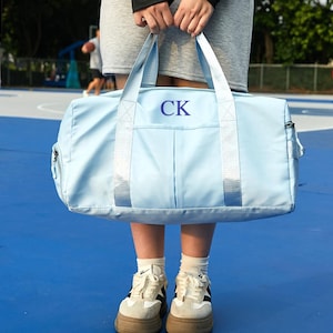 Op de afbeelding: Een lichtblauwe duffeltas met de initialen "CK" in marineblauw. De tas heeft een rechthoekige vorm, een voorvak en twee handvatten. De tas wordt vastgehouden door een persoon die een grijze rok en witte sneakers draagt.