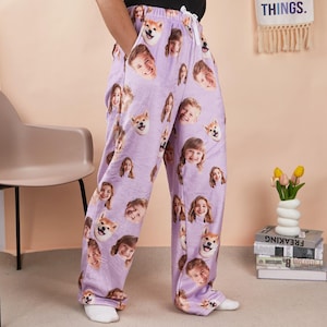 Puede incluir: Pantalones de pijama color lavanda con un patrón repetido de caras sonrientes y caras de perros Shiba Inu. Los pantalones tienen cintura con cordón y bolsillos. La palabra "THINGS" es visible en el fondo.