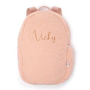 May include: A pale pink, fluffy backpack with the name "Vicky" embroidered in gold thread. The backpack features a rounded design, a small side pocket, and adjustable shoulder straps. The material has a soft, textured finish.