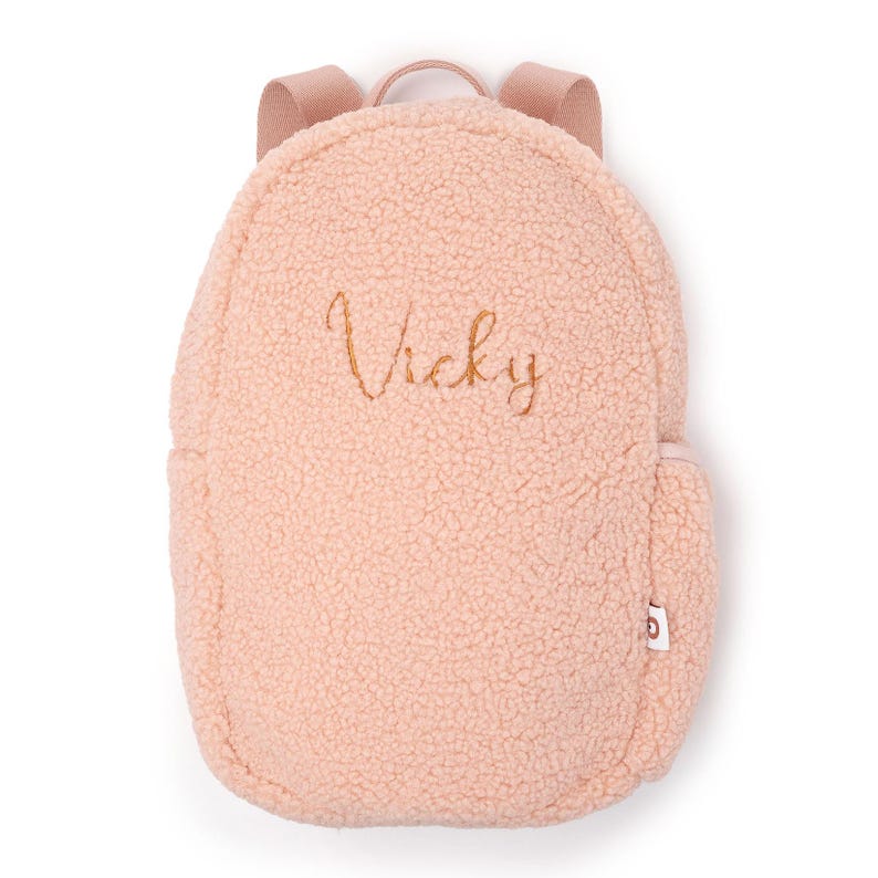 May include: A pale pink, fluffy backpack with the name "Vicky" embroidered in gold thread. The backpack features a rounded design, a small side pocket, and adjustable shoulder straps. The material has a soft, textured finish.