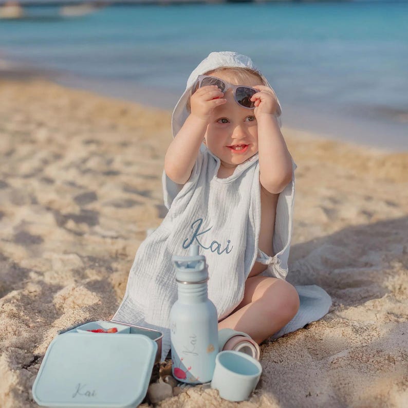 May include: A child wearing a light blue hooded beach cover-up and sunglasses, sitting on the sand. A matching light blue lunchbox, water bottle, and cup are nearby. The name "Kai" is embroidered on the cover-up and lunchbox.