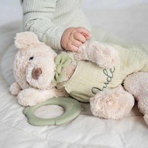 May include: A soft beige teddy bear toy with a green bow and a green outfit. The bear has a brown nose and black eyes. A green teething ring is next to the bear. The name "Jude" is embroidered on the outfit.