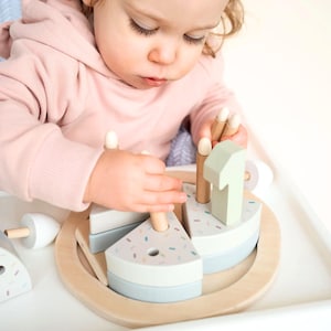 May include: A wooden toy cake with a light blue and white design, featuring a number 1 and wooden candle pegs. The cake sits on a wooden tray and is decorated with colourful sprinkles. A child is playing with the toy.