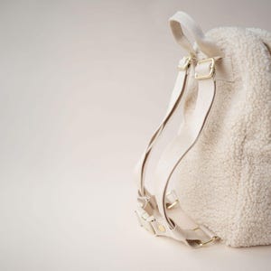 May include: Cream-coloured backpack with a fluffy, textured exterior. The bag features adjustable cream-coloured straps and gold-coloured metal hardware. The backpack has a top handle and a zipped closure.