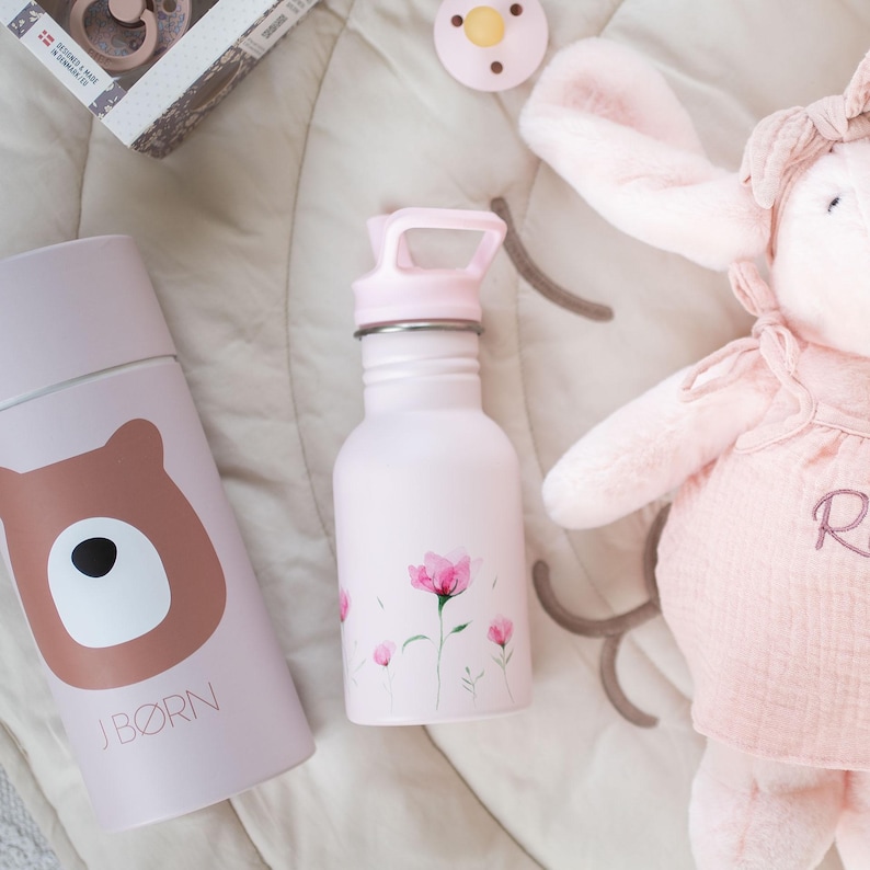 May include: A light pink water bottle with a floral design and a matching pink lid. Next to it is a light pink insulated bottle with a brown bear graphic and the text "J B&Oslash;RN". A pink pacifier and a plush bunny are also in the image.