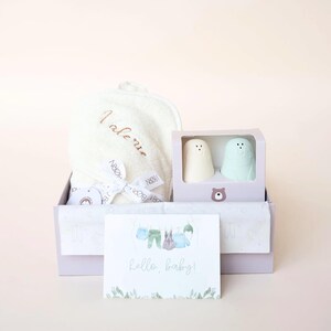 Puede incluir: Un set de regalo para bebé en una caja lila. El set incluye una toalla con capucha blanca, dos juguetes de baño con forma de oso y una tarjeta que dice "hello, baby!". La tarjeta tiene una ilustración de acuarela de ropa de bebé.