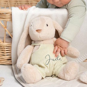 May include: A plush beige bunny wearing light green overalls with the name "Jude" embroidered in cursive. The bunny has long floppy ears and is sitting on a white blanket. A baby's hand is touching the bunny.