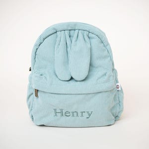 May include: A pale blue corduroy backpack with a bunny ear design. The backpack features a front pocket and the name "Henry" embroidered on the front. It has a zip closure and a small tag on the side.