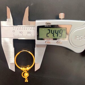 May include: A gold hoop earring with a dangling charm. The earring is being measured with a caliper, which reads 24.49 mm.