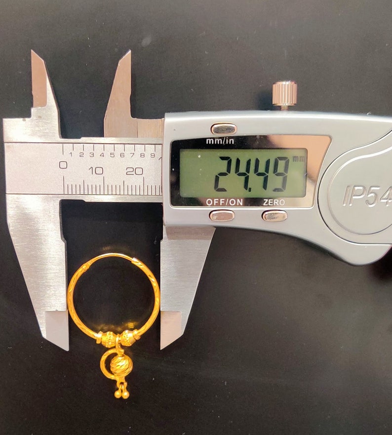 May include: A gold hoop earring with a dangling charm. The earring is being measured with a caliper, which reads 24.49 mm.