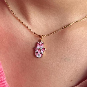 Könnte beinhalten: Eine goldene Kettenkette mit einem Anhänger in Form einer Hamsa-Hand aus rosa und weißem Emaille. Die Hamsa-Hand ist ein Symbol für Schutz und Glück.