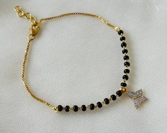 Könnte beinhalten: Ein goldenes Kettenarmband mit schwarzen Perlen und einem Schmetterlingsanhänger. Der Schmetterlingsanhänger ist mit kleinen, klaren Steinen verziert.