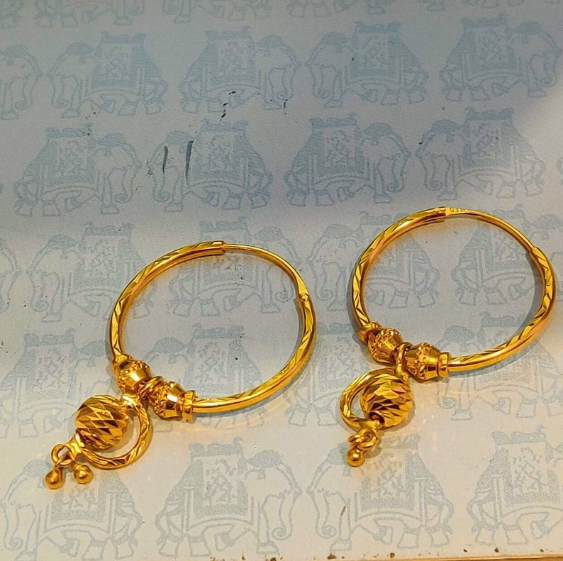 May include: A pair of gold hoop earrings with a decorative charm hanging from each hoop. The charms are round and have a textured surface.