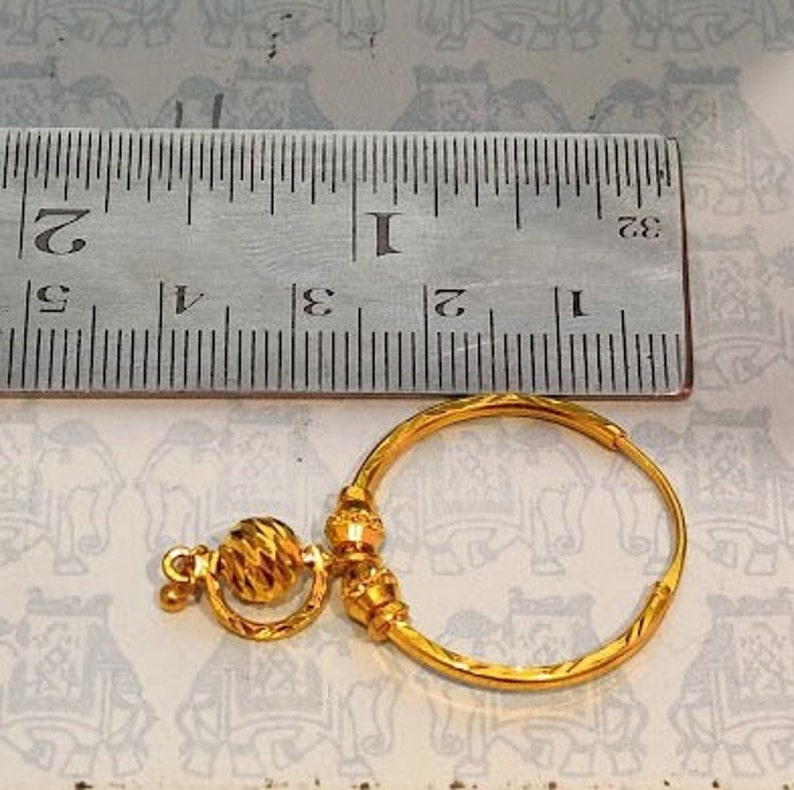 May include: A gold hoop earring with a dangling ball charm. The earring is measured against a ruler.