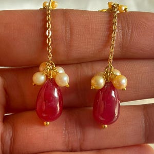 May include: A pair of dangle earrings featuring a red teardrop-shaped gemstone and a cluster of white pearls suspended from a gold chain.
