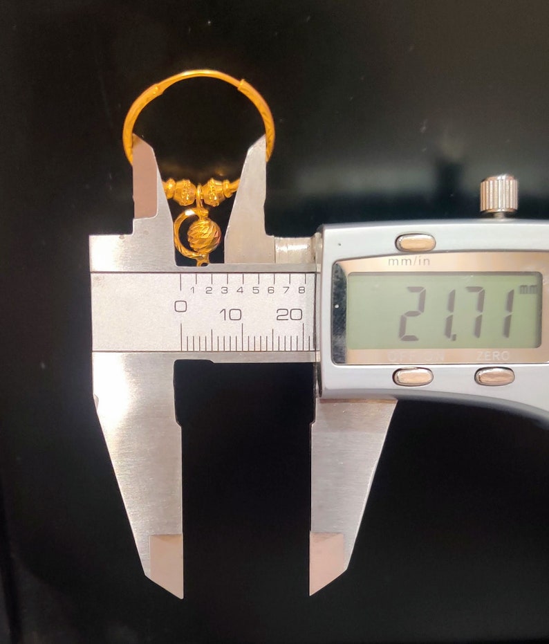 May include: A gold hoop earring with a small, round, decorative charm hanging from it. The earring is measured with a caliper, showing a measurement of 21.71 mm.