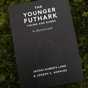 May include: A black book titled "The Younger Futhark Viking Age Runes" with white text and an illustration of a rune stone. The book is lying on a bed of green moss. The author's names are also listed.
