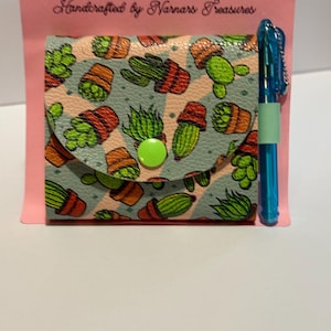 以下が含まれることがあります： サボテン柄の付箋カバー、緑色のボタン、青いペン。カバーは水色で、オレンジ色の鉢に入った緑色のサボテンの繰り返し模様です。「POST-IT-NOTE COVER」と「Handcrafted by Narnars Treasures」の文字が見えます。