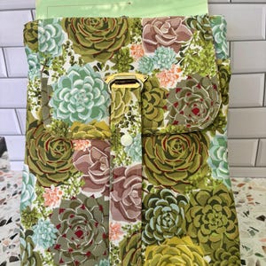 May include: A green, white, and pink floral fabric zippered pouch with a gold buckle closure. The pouch is designed to hold a tablet or other flat items.