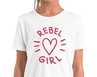 May include: White t-shirt with the words "REBEL GIRL" in red, along with a red heart graphic. The shirt is short-sleeved and appears to be made of a soft material. The design is centered on the front of the shirt.