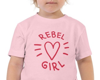 May include: A light pink toddler t-shirt with the words "REBEL GIRL" in red, surrounding a red heart graphic. The shirt is short-sleeved and made of a soft material.