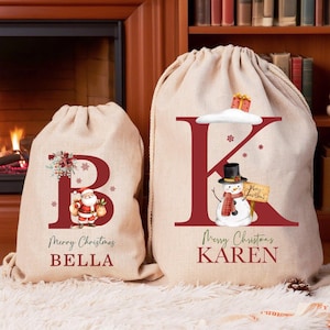 Könnte beinhalten: Zwei beige Weihnachtstüten mit Kordelzugverschlüssen. Eine Tasche zeigt den roten Buchstaben "B" mit einer Weihnachtsmann-Grafik und dem Namen "BELLA". Die andere Tasche hat ein rotes "K" mit einer Schneemann-Grafik und dem Namen "KAREN". Beide Taschen sagen "Frohe Weihnachten".