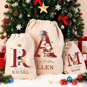 Könnte beinhalten: Drei beige Geschenksäcke aus Jute mit Kordelzug, verziert mit großen kastanienbraunen Buchstaben R, A und M. Die Säcke zeigen Illustrationen eines Schneemanns, des Weihnachtsmanns und floraler Akzente sowie die Namen Rachel, Allen und Mark.