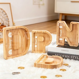 Könnte beinhalten: Holzsparbüchsen in Buchstabenform, die den Namen "DPN" bilden. Die Buchstaben "D", "P" und "N" sind zu sehen. Jede Bank hat eine transparente Vorderseite und ist mit einem Namen und einer Grafik verziert. Münzen liegen verstreut.