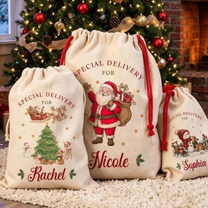 May include: Three beige canvas drawstring gift sacks with red rope cords. The sacks feature printed Christmas graphics and the names Rachel, Nicole, and Sophia. Each bag is labeled with the text Special Delivery For followed by a name.