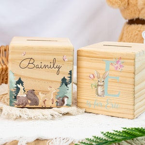 May include: Two wooden money boxes with personalised names. One box features a woodland scene with a bear, deer, and squirrel. The other box has a light blue letter E with a bunny holding a pink flower. The text "E is for Eric" is written below the letter.