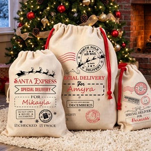 May include: Three beige drawstring canvas sacks with red cords, featuring printed North Pole air mail graphics, Santa Express branding, and personalized names Mikayla, Amyra, and Princess, with holiday-themed stamps and text details.