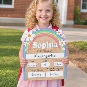 K&ouml;nnte beinhalten: Ein Holzschild mit Regenbogen- und G&auml;nsebl&uuml;mchen-Design, personalisiert mit dem Namen "Sophia" und Details f&uuml;r ihren ersten Kindergartentag. Das Schild enth&auml;lt Datum, Alter, Gr&ouml;&szlig;e (110cm) und Interessen: Zeichnen und Lehrerin.