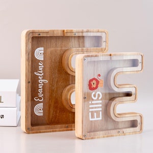 Może przedstawiać: Dwie drewniane skarbonki w kształcie liter E i O. Skarbonka E ma grafikę lwa i napis "Ellis". Skarbonka O ma grafikę tęczy i napis "Evangeline".