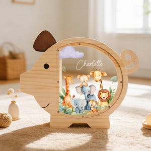 Könnte beinhalten: Holzsparschwein in Schweineform mit einem transparenten kreisförmigen Fenster, das eine Dschungelszene mit Zeichentieren zeigt. Der Name "Charlotte" steht auf dem Fenster. Die Spardose ist aus hellem Holz gefertigt.