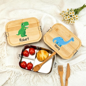 Puede incluir: Dos fiambreras rectangulares con tapas de bambú que presentan un T-Rex verde con el nombre Robert y un dinosaurio azul de cuello largo con el nombre Henry. Un recipiente metálico abierto contiene cuatro tomates cherry, un panecillo y una cuchara de madera.