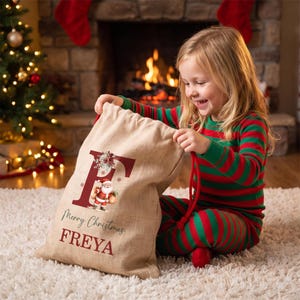 Puede incluir: Un saco de regalo de Navidad de arpillera beige con una letra F grande de color rojo oscuro, una peque&ntilde;a ilustraci&oacute;n de Santa Claus y el texto Merry Christmas FREYA impreso en tinta roja oscura y verde.