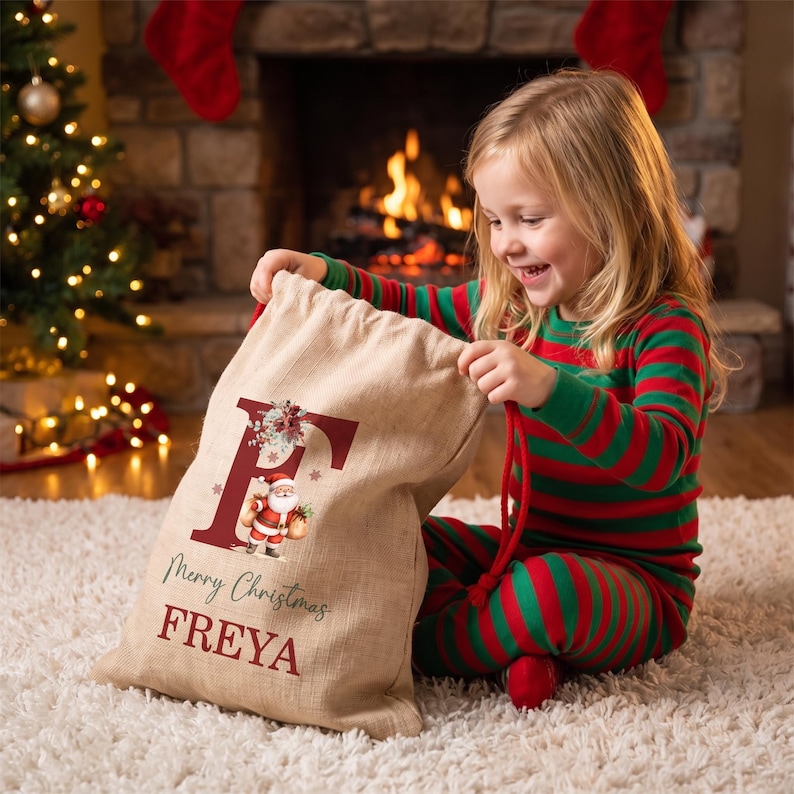 Puede incluir: Un saco de regalo de Navidad de arpillera beige con una letra F grande de color rojo oscuro, una peque&ntilde;a ilustraci&oacute;n de Santa Claus y el texto Merry Christmas FREYA impreso en tinta roja oscura y verde.