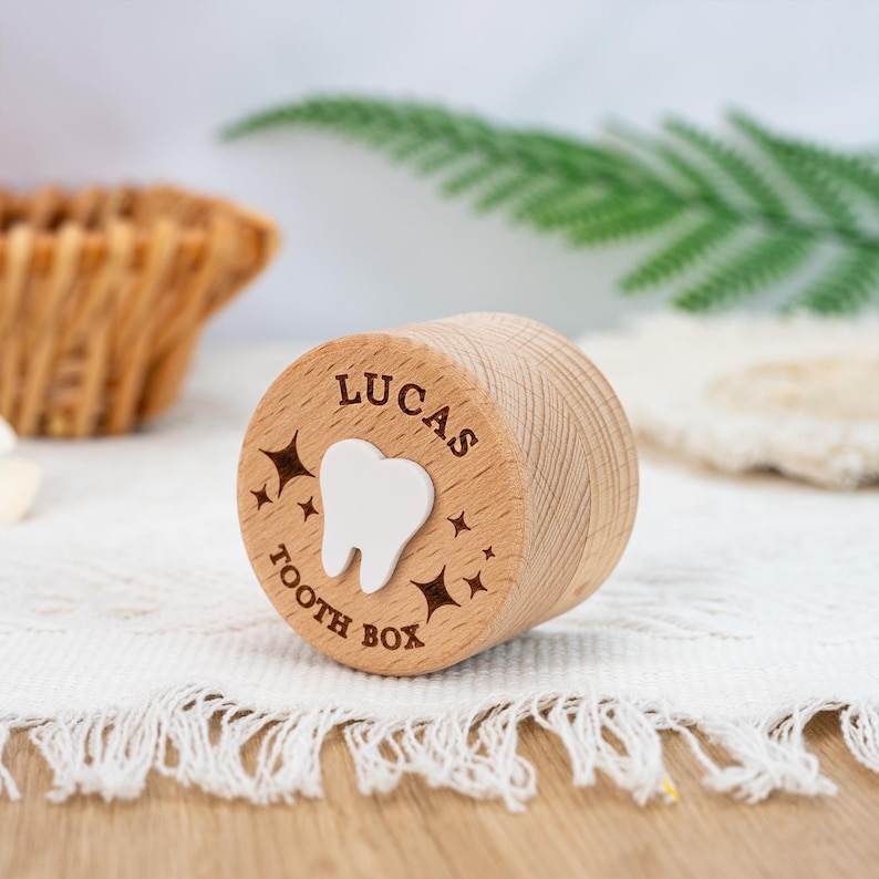 May include: A cylindrical light brown wooden tooth box featuring the engraved name LUCAS and the text TOOTH BOX. A white tooth icon is centred on the lid, surrounded by small engraved star shapes. The wood grain is visible on the side surface.