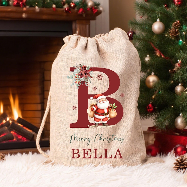 Puede incluir: Una bolsa de regalo de arpillera beige con cord&oacute;n, que presenta una letra B grande de color rojo oscuro, una ilustraci&oacute;n de Santa Claus y el texto Merry Christmas BELLA impreso en letras rojas y verdes en la parte frontal.