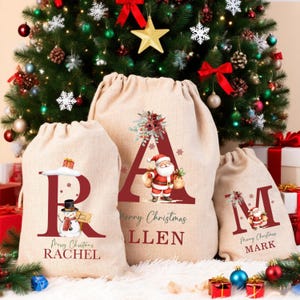 May include: Three beige Christmas gift bags with red lettering and festive illustrations. The bags feature the letters "R", "A", and "M", with the names "Rachel", "Allen", and "Mark" below. A decorated Christmas tree is in the background.