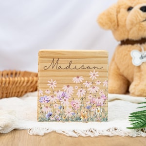 以下が含まれることがあります： 「Madison」と筆記体で書かれた木製のブロック。ピンク、紫、白の花柄で装飾されています。白いレースの布の上に置かれています。