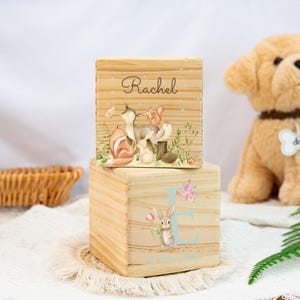 May include: Two wooden blocks with a woodland theme. The top block features a watercolour illustration of a fox, deer, squirrel, and rabbit. The text "Rachel" is printed on the block. The bottom block features a watercolour illustration of a rabbit and the letter "E" in a light blue circle. The text "Ellie Lou" is printed on the block.