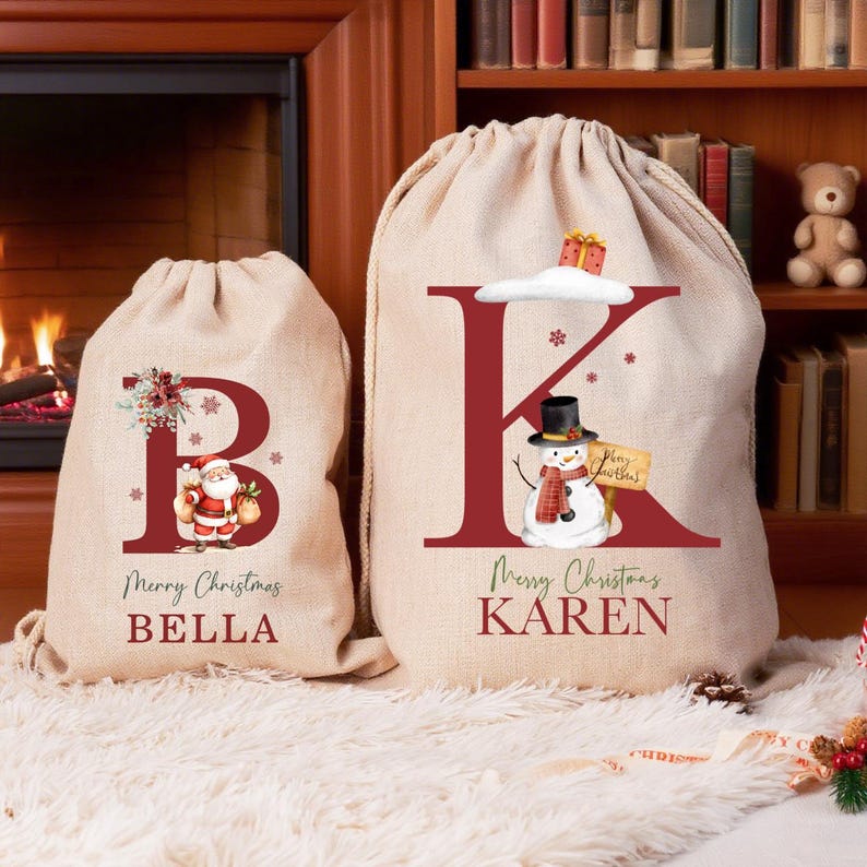 Puede incluir: Dos bolsas de lona beige con cord&oacute;n y letras rojas. La bolsa m&aacute;s peque&ntilde;a presenta una letra B grande, una ilustraci&oacute;n de Santa Claus y el nombre BELLA. La bolsa m&aacute;s grande presenta una letra K grande, un mu&ntilde;eco de nieve y el nombre KAREN.